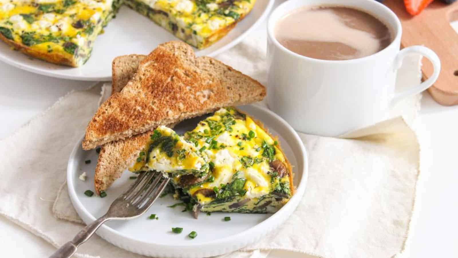 A slice of egg frittata with spinach, mushrooms, and goat cheese sitting on a white plate with two triangle slices of toast and a coffee mug behind it.