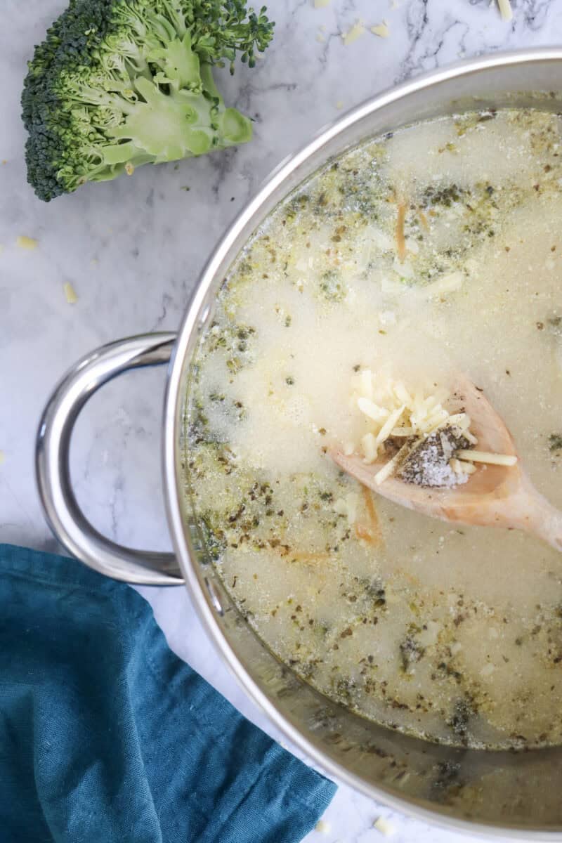 Close-up image of shredded cheddar, salt, and pepper in a wooden laddle being added to the soup.