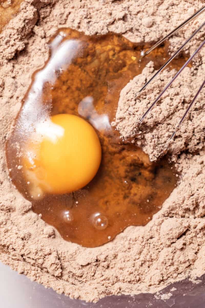 Close-up image of an egg on top of brown powder with a whisk dipped into the mixture.