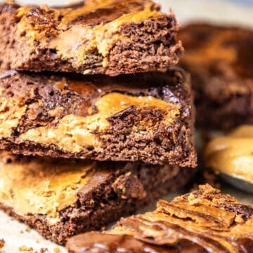 "Close-up image of a batch of peanut butter brownies with a stack of three in the middle, accompanied by a spoonful of peanut butter beside it.