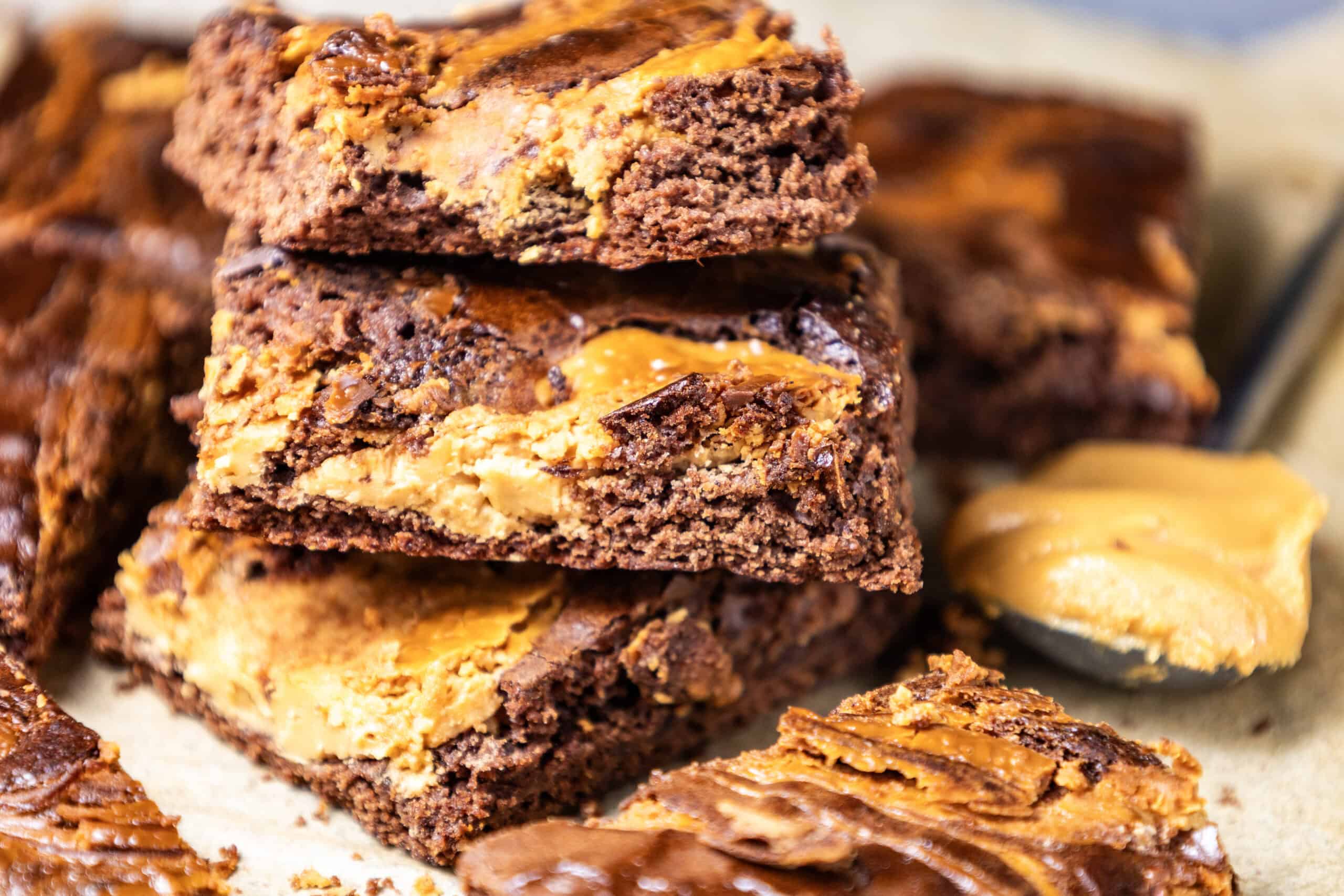 "Close-up image of a batch of peanut butter brownies with a stack of three in the middle, accompanied by a spoonful of peanut butter beside it.