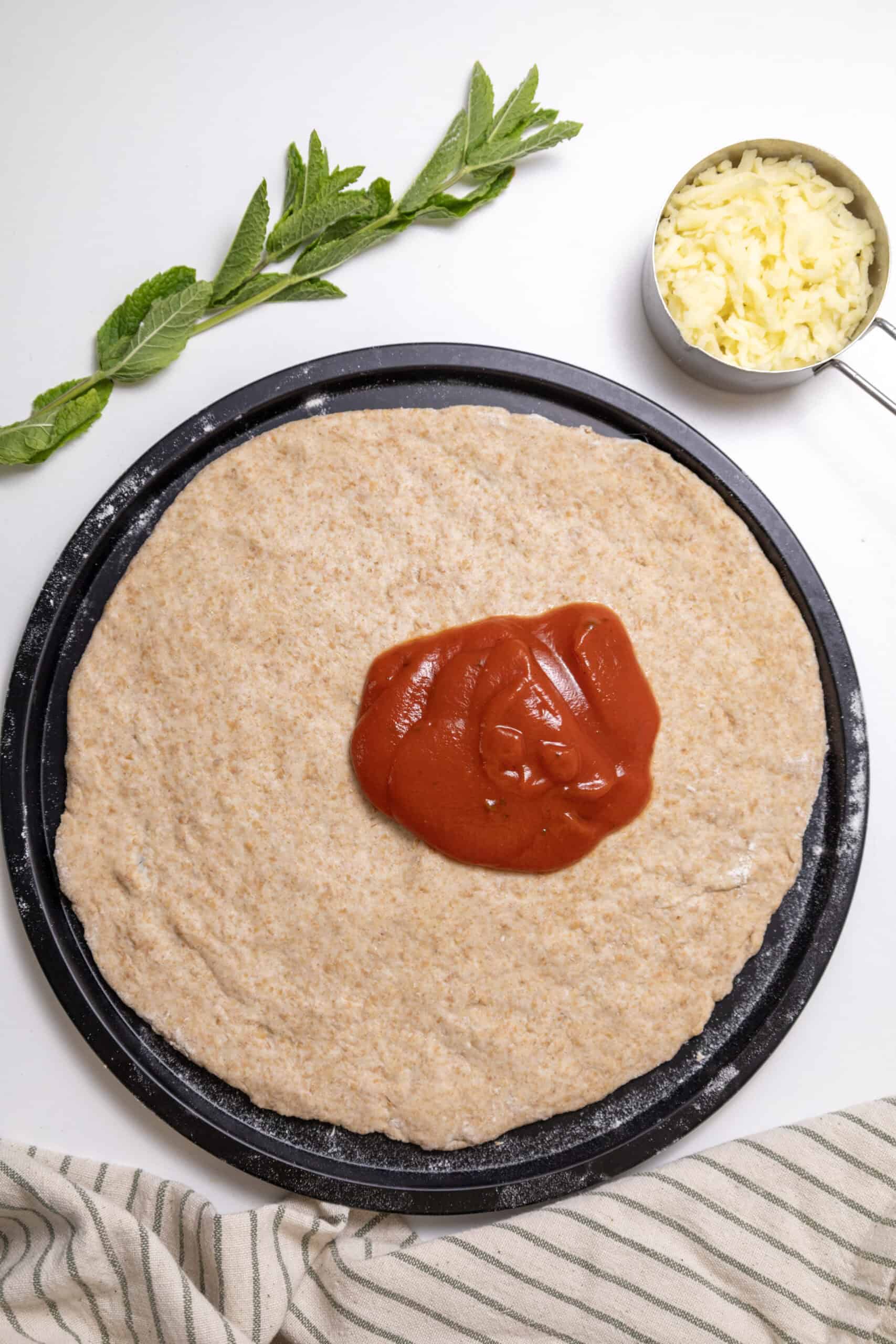 Pizza dough in a pizza pan with sauce on top, with cheese in the background.