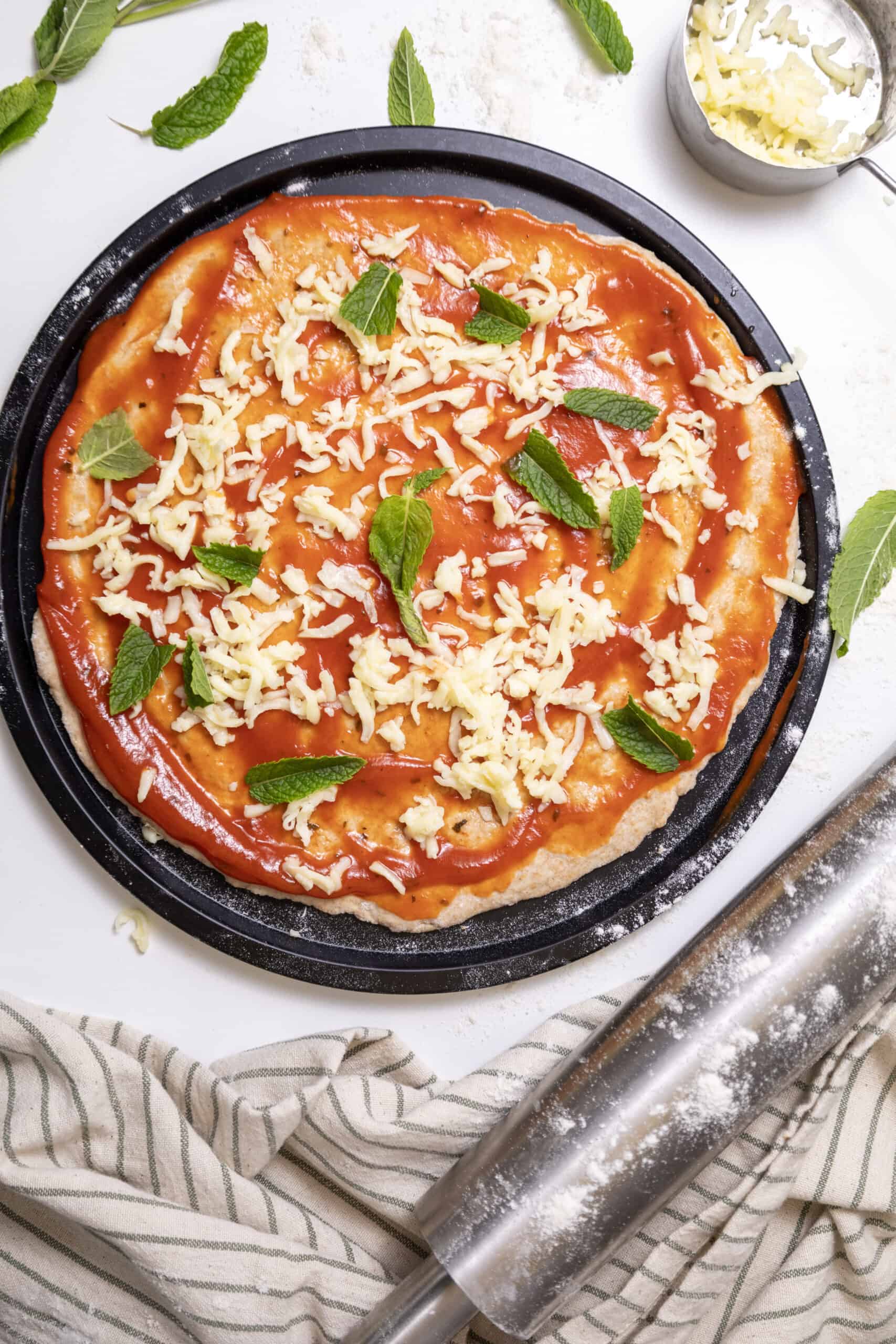 Pizza dough in a pizza pan with sauce, grated cheese, and mint leaves spread on top.