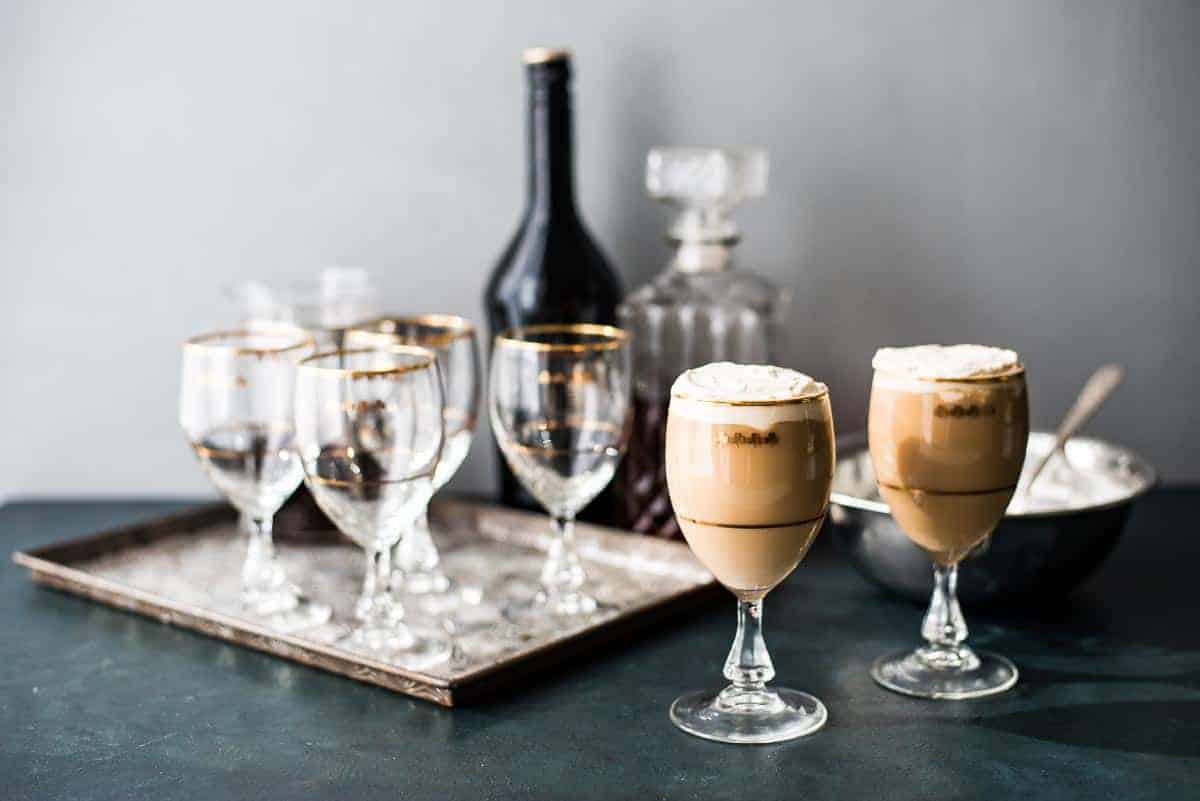 Two glasses of Irish Cream coffee in front of a tray of festive glass and a bar set up for making more.