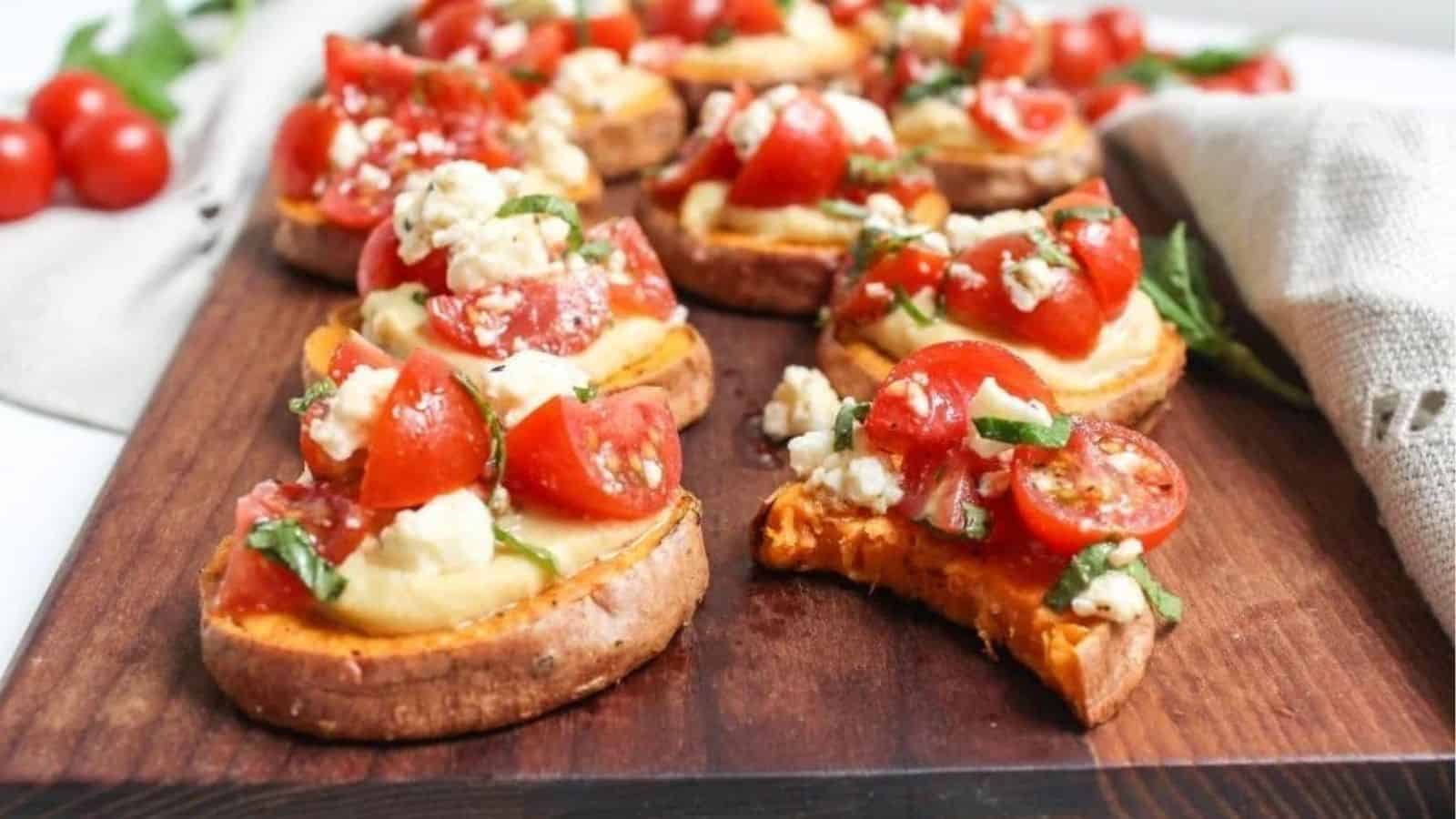 A wooden board with sweet potato rounds topped with hummus and diced tomatoes.