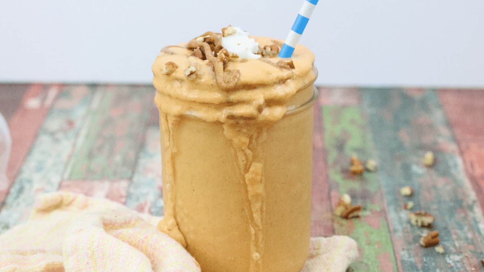 Mason jar with light orange pumpkin smoothie tipped with coconut whip cream, nuts and a blue straw spilling down the side.