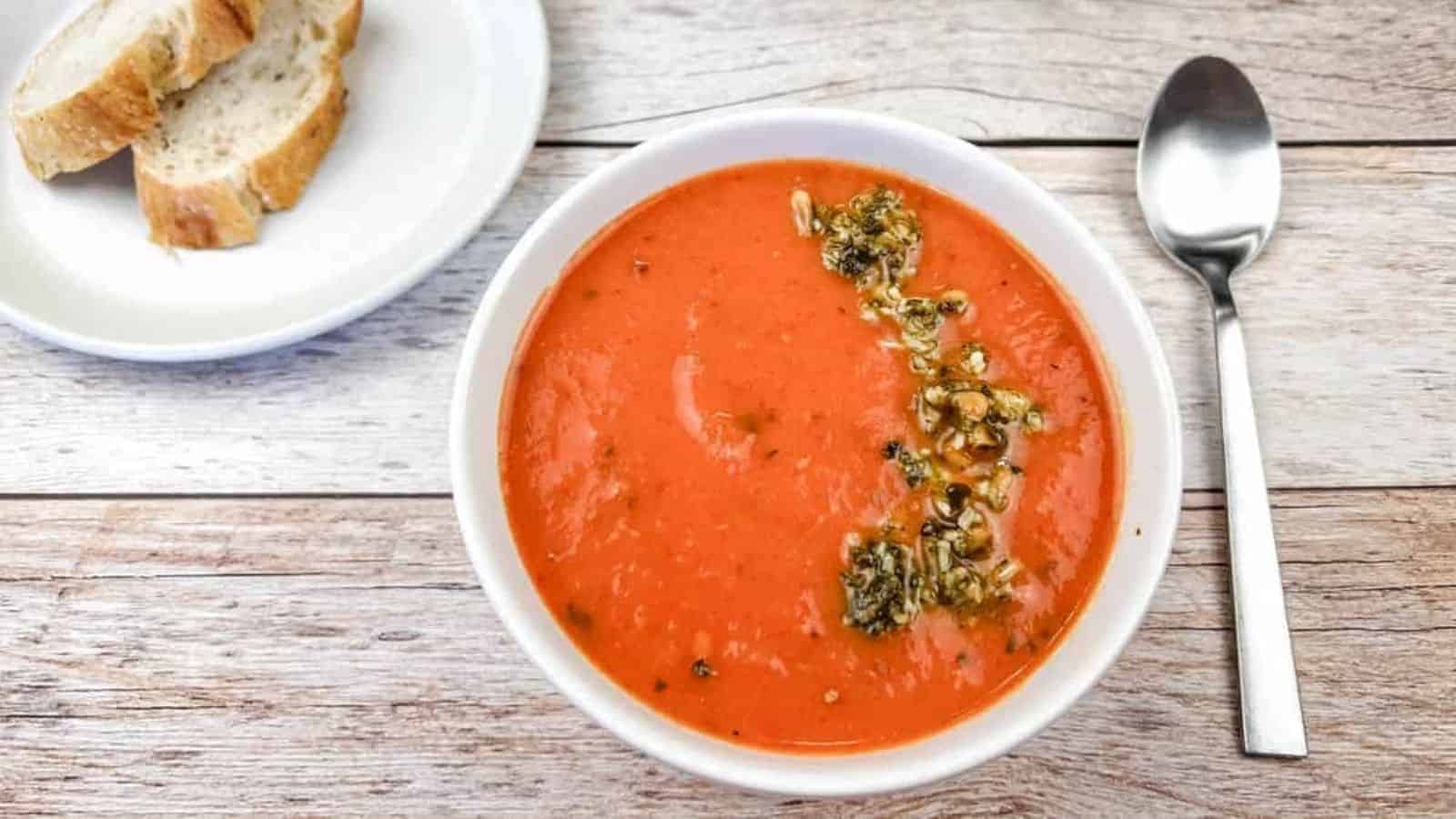 A bowl of red pepper and tomato soup next to a plate of bread and a spoon.