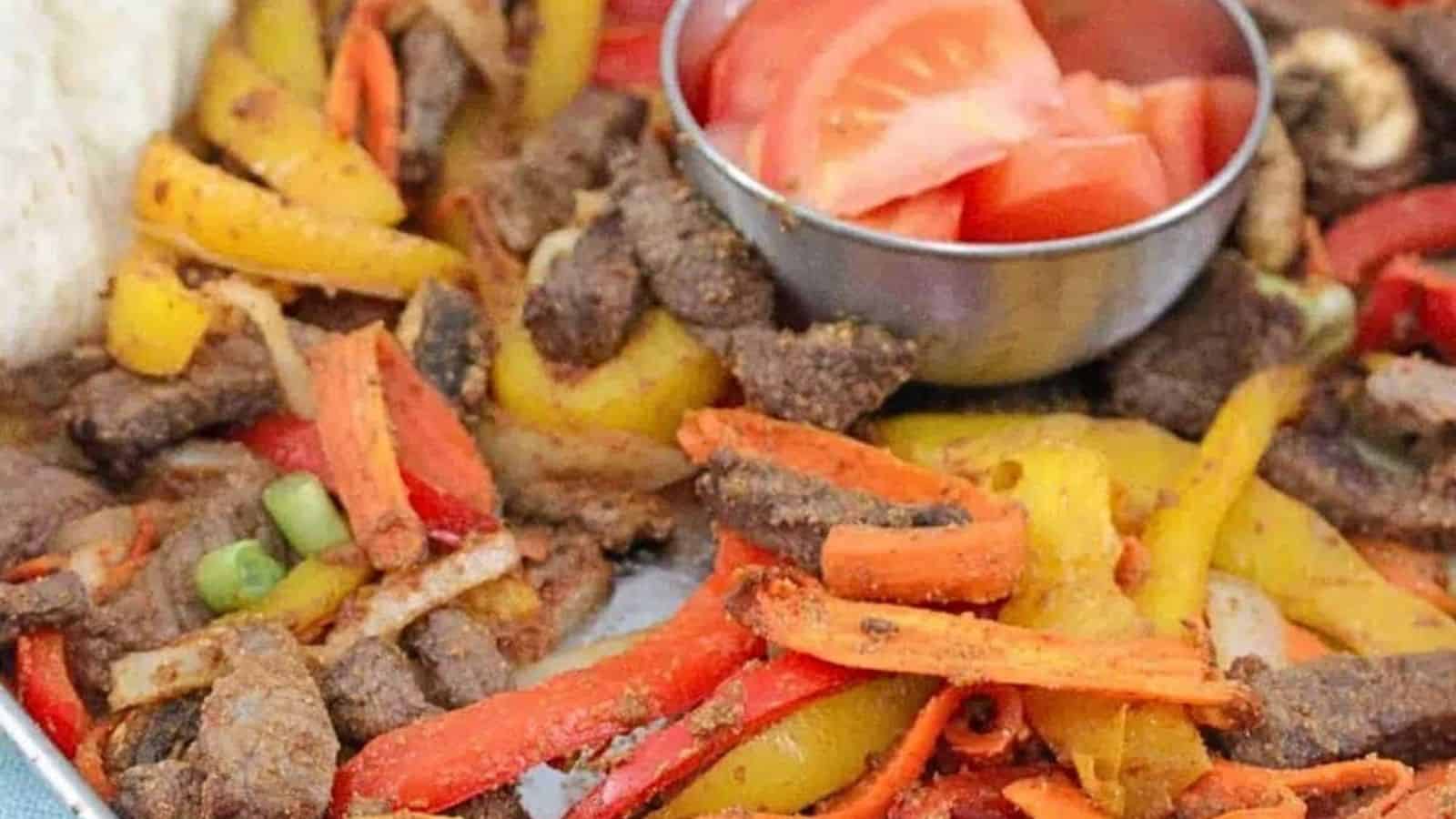 Baking tray with cooked steak, bell peppers, and veggies with a steel bowl of fresh tomatoes in the middle and flour tortillas folded to the side.