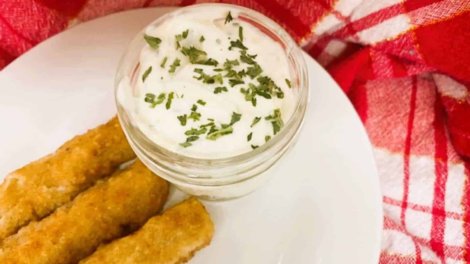 Tartar sauce in clear jar on white plate of fish sticks.
