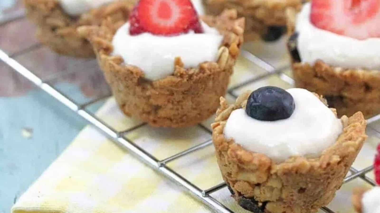 Mini granola cups topped with yogurt and blueberries and strawberries on a cooling rack over a yellow linen.