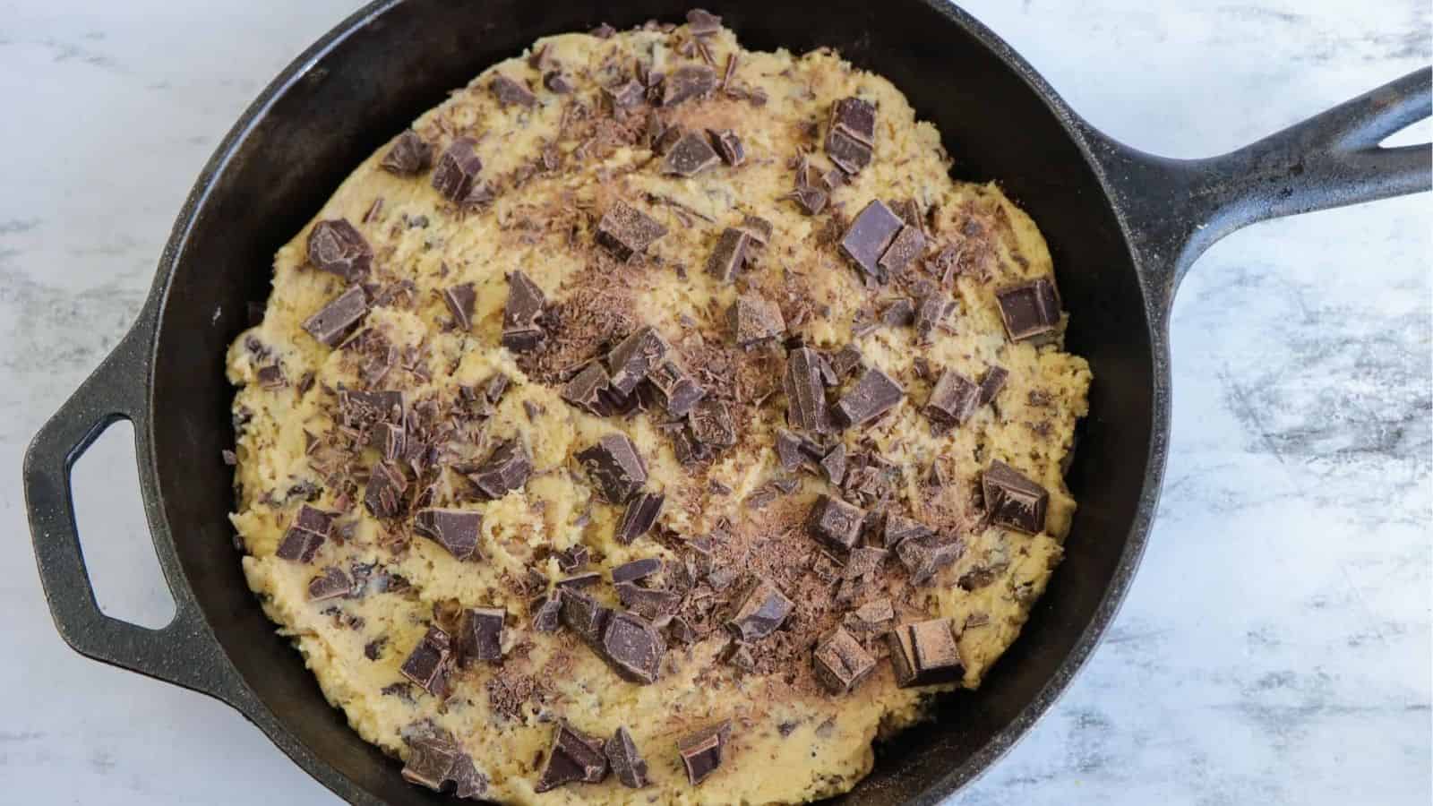 Chocolate chip skillet cookie batter topped with chopped chocolate bar in a skillet.