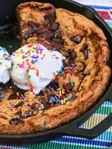 Close-up image of a chocolate chip skillet cookie topped with two scoops of ice cream and sprinkles, with one slice missing.