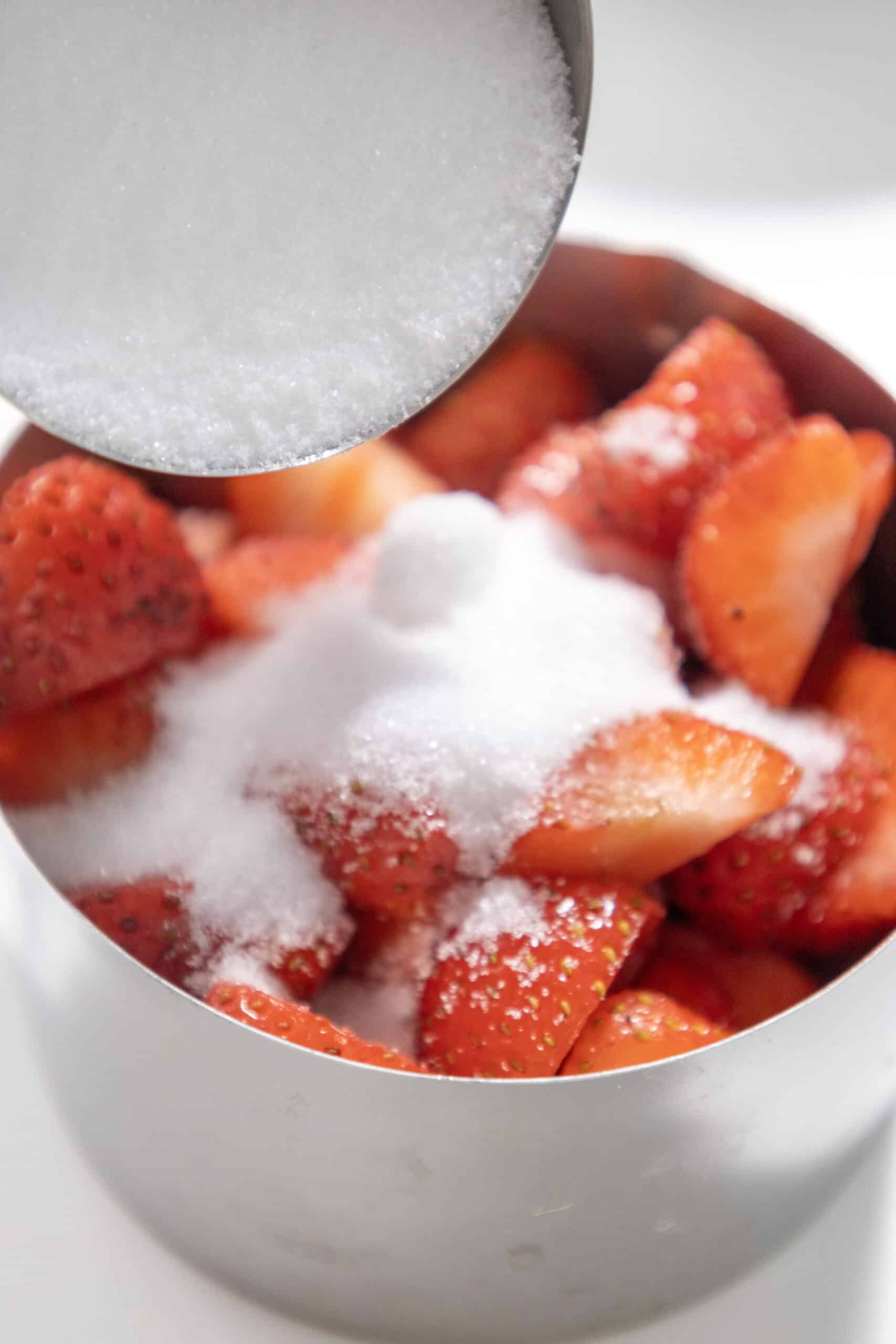 Close-up image of sugar being poured over diced strawberries in a bowl.