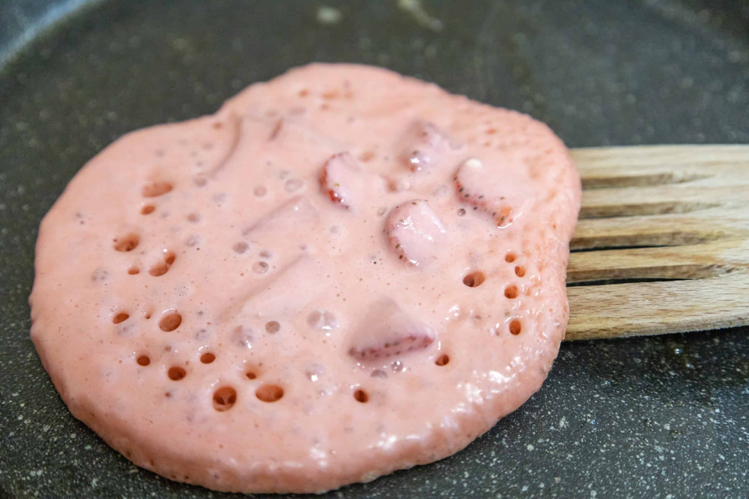 Strawberry Delight Pancakes resting on a wooden spatula, poised above a skillet, ready to be flipped.