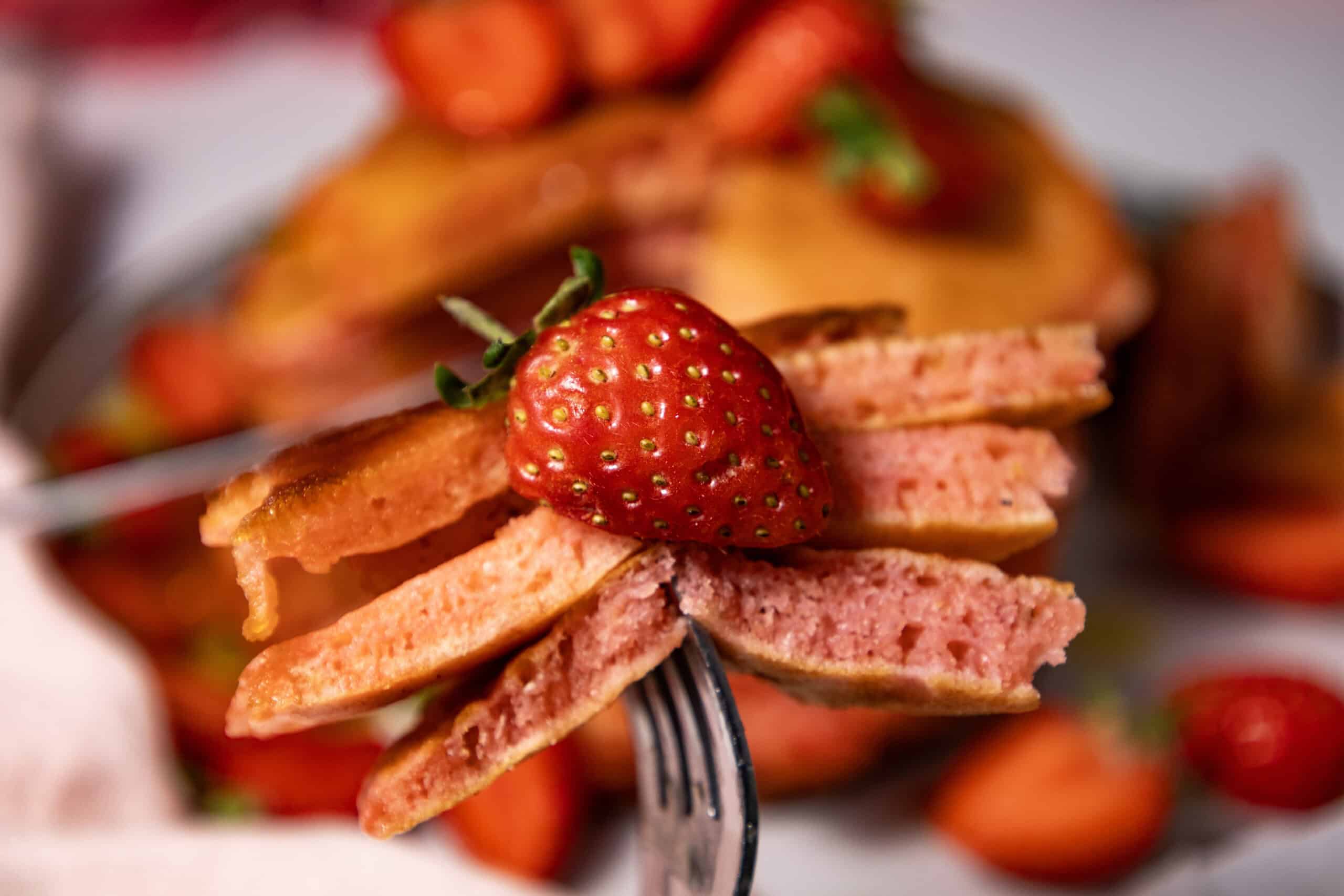 Close-up image of a fork holding a portion of a stack of Strawberry Delight Pancakes with sliced strawberry.