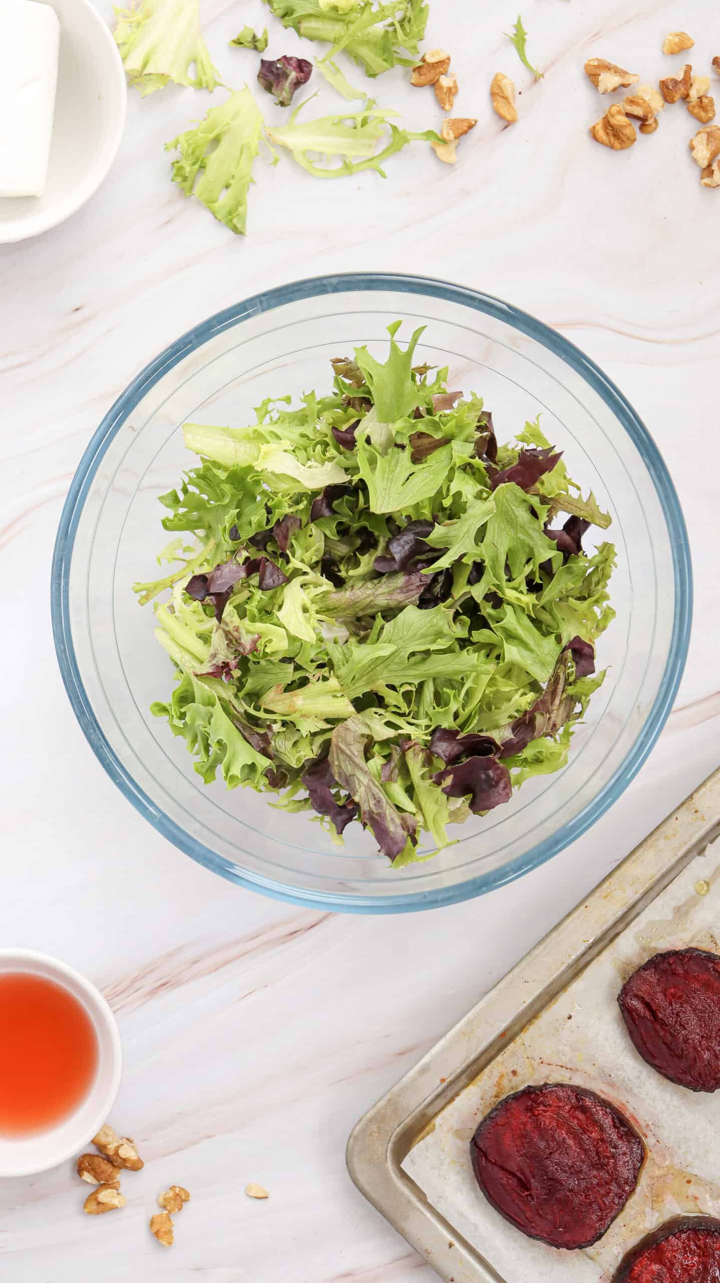 Overhead image of mixed greens in a bowl, surrounded by roasted beetroot slices on a sheet pan, goat cheese, chopped walnuts and red vinaigrette in the background.