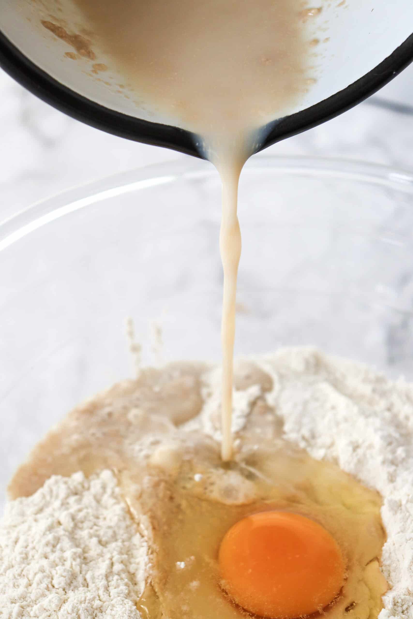 A large mixing bowl filled with flour, salt, and yeast, with an egg and the milk mixture being added.