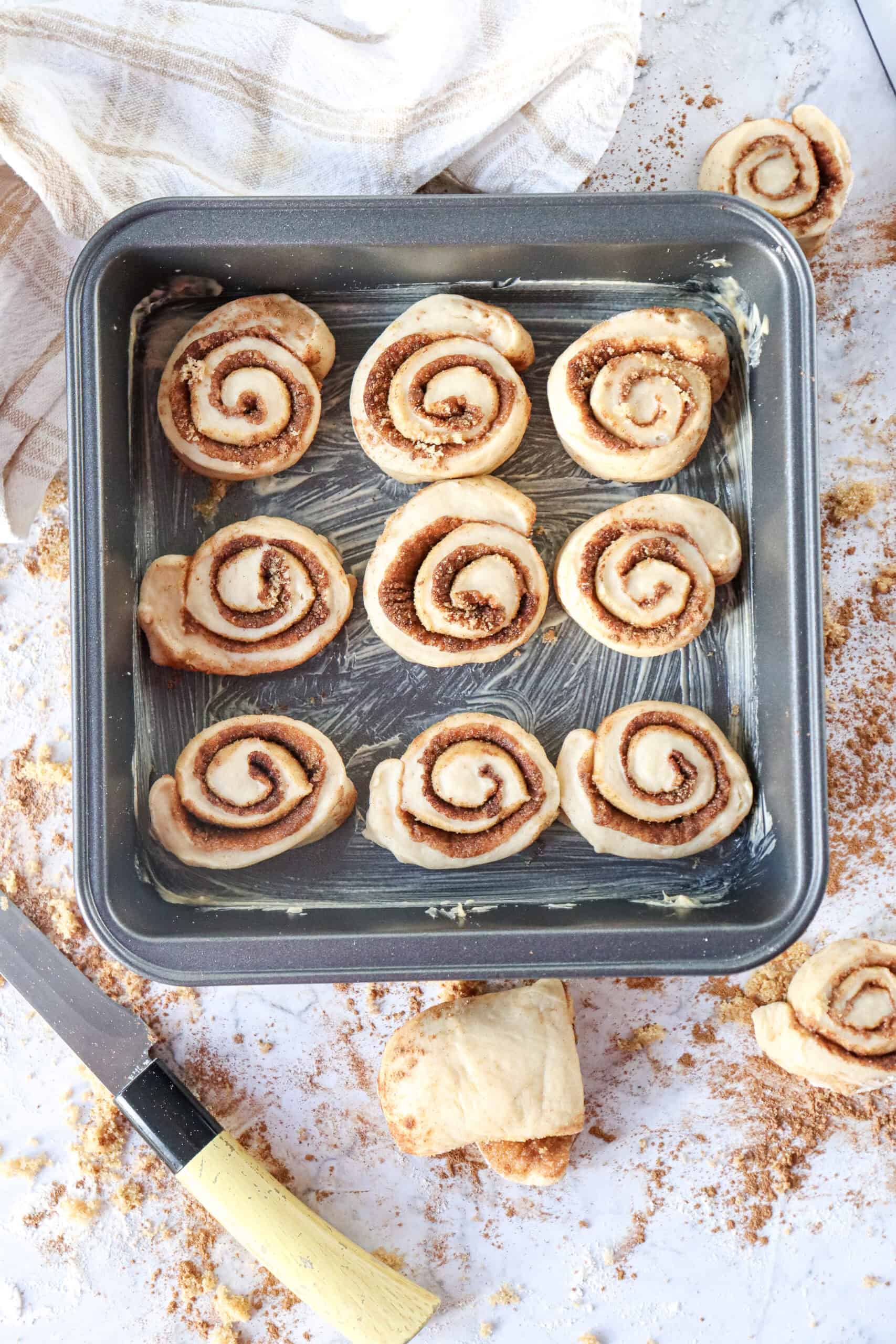 Cinnamon rolls arranged in a greased square or rectangular baking dish, with some space left between them.