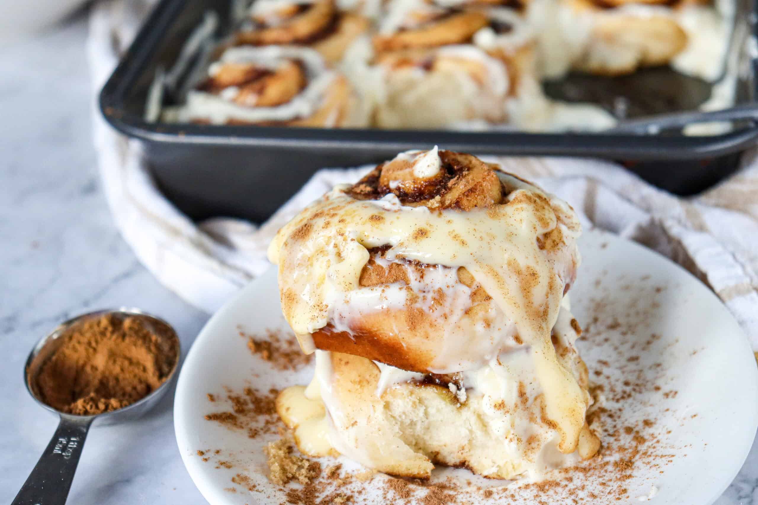 A stack of two cinnamon roll pieces on a plate, with the square pan of remaining rolls in the background.