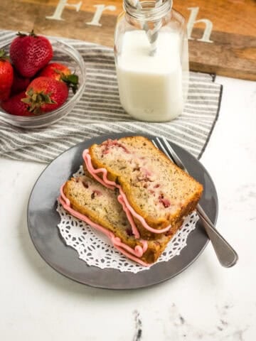 Close-up image of two slices of strawberry banana bread on a plate with a fork. A small glass bowl holds fresh strawberries, and a glass of milk is visible in the background.