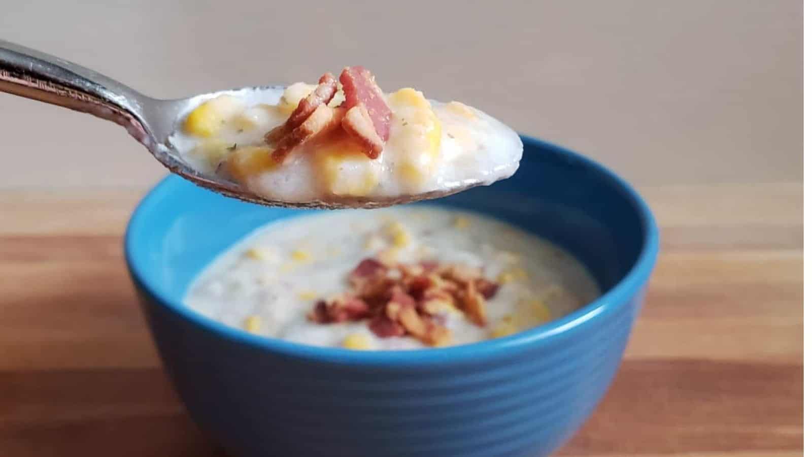 Spoon holding potato and corn chowder with bacon with the full bowl of soup behind it.