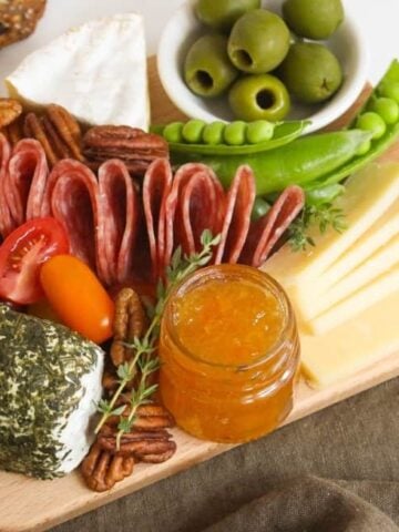 A small wooden board topped with cured meat, sliced cheese, dips, vegetables, nuts, and fruits.