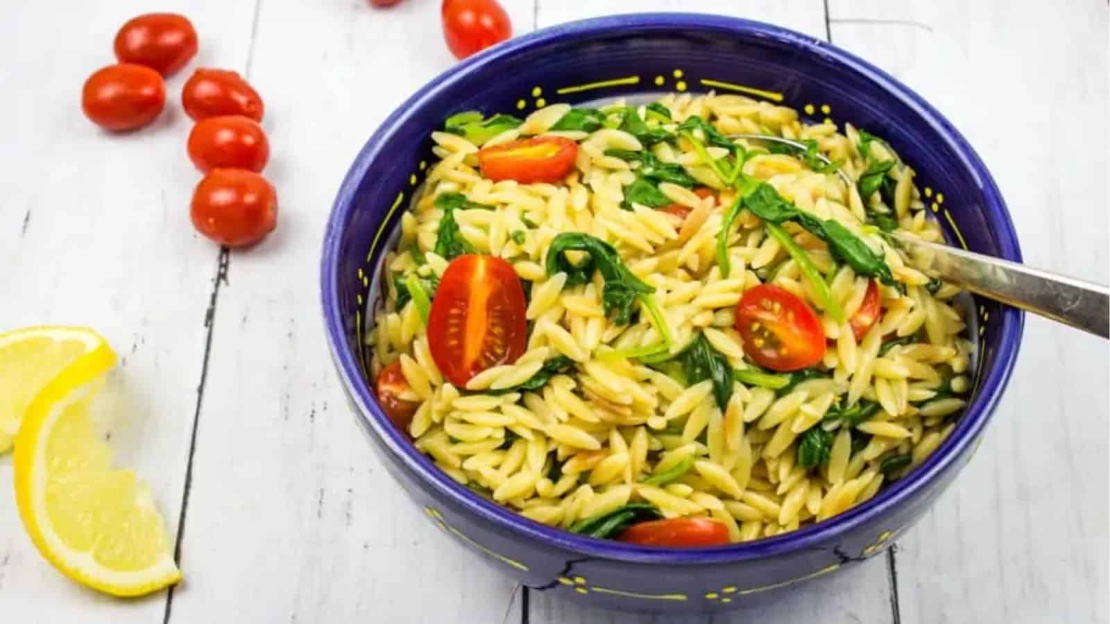 A bowl of orzo pasta with spinach and cherry tomatoes on a wooden table, with a spoon inside and lemon wedges nearby.