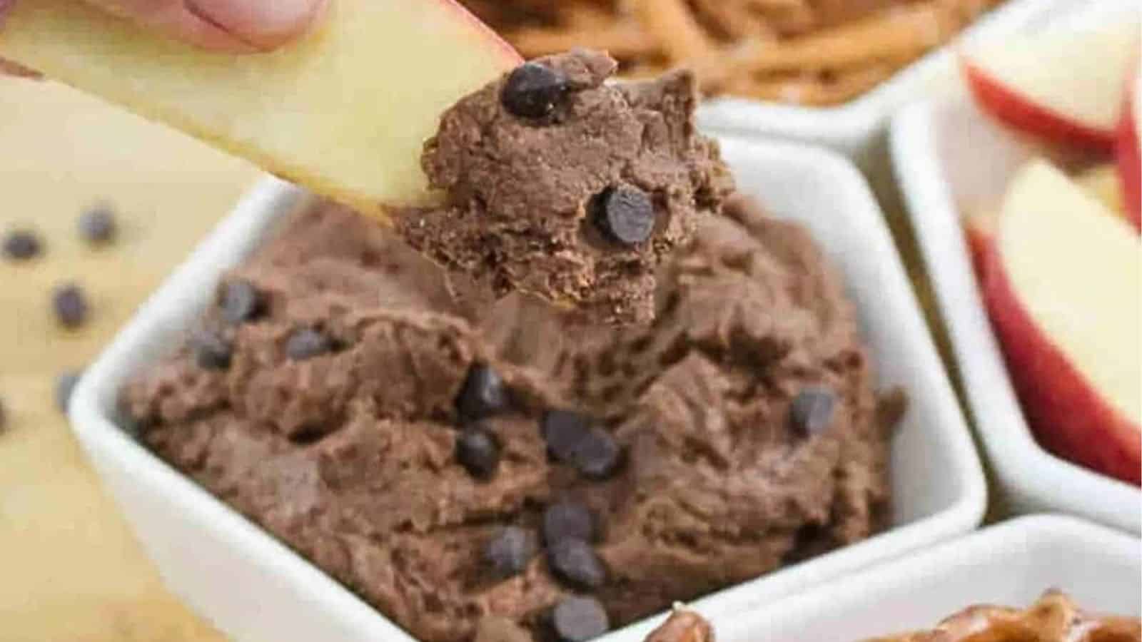 Wooden serving platter with edible brownie batter in a white ramekin surrounded by pretzels and apples and a hand dipping an apple into the brownie batter dip.