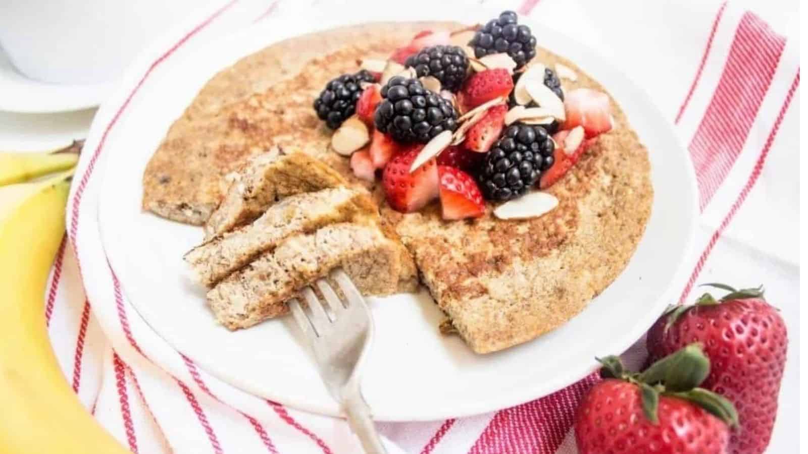 A banana pancake on a plate topped with berries and sliced almonds, with a few bites cut out on a fork.