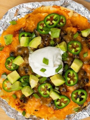 An overview of air fryer nachos on a foil on top of a wooden board with a jalapeño and some slices of lime on its sides.