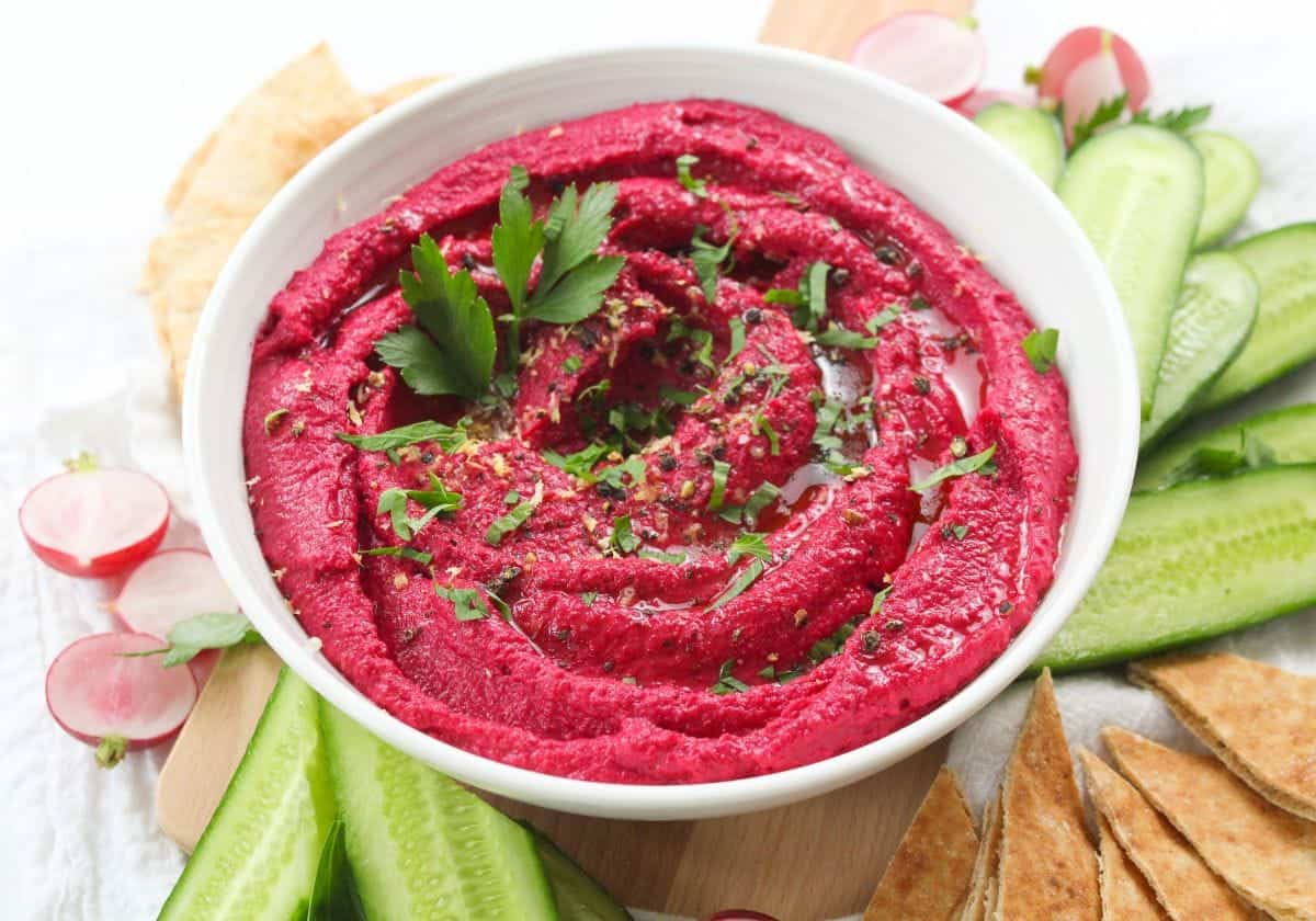A bowl of pink beetroot hummus with cucumbers, radishes, and pita bread.