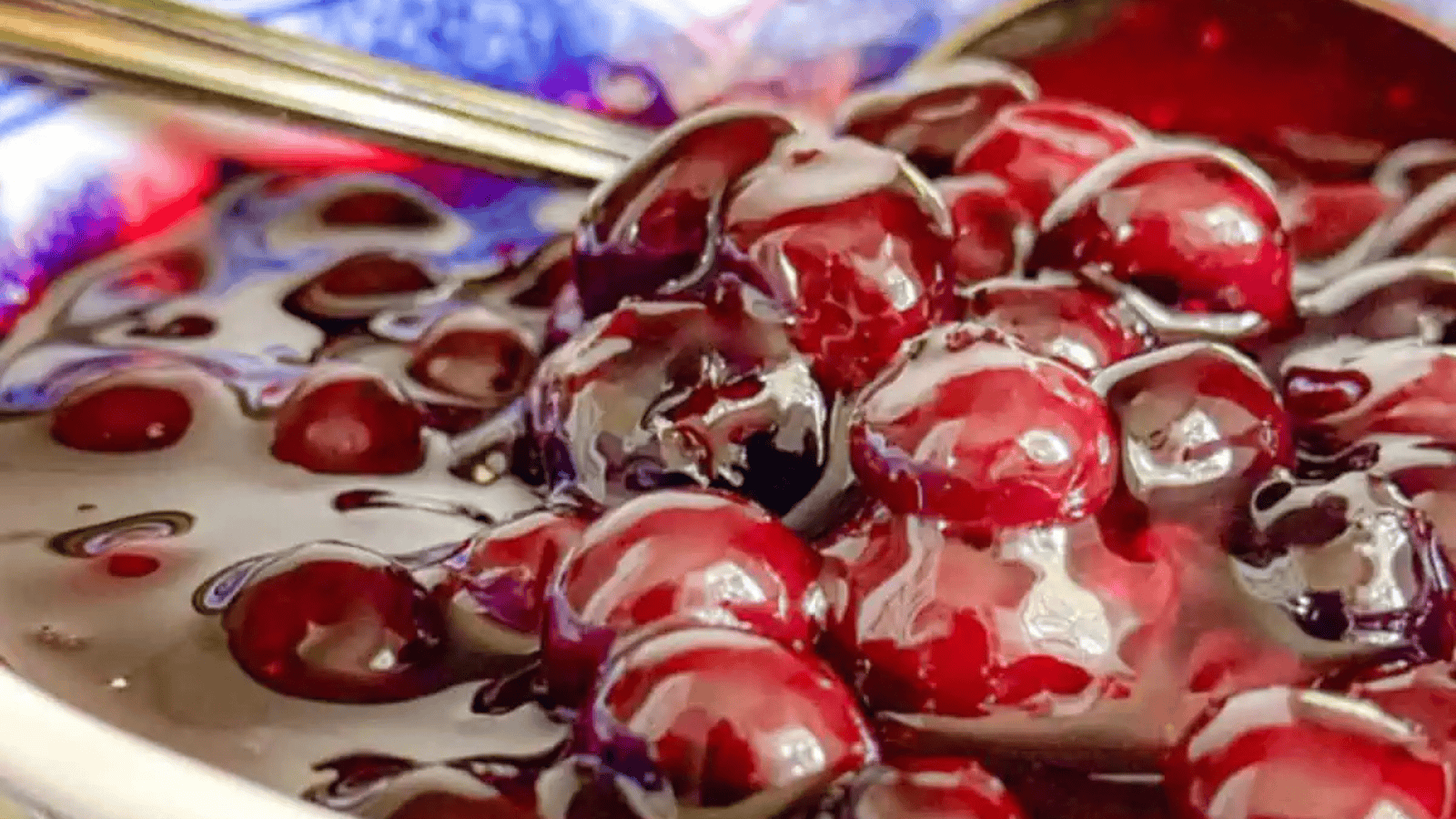 A close-up shot of a bowl filled with glistening cherry compote. The cherries are plump and coated in a thick, glossy syrup, with a spoon partially visible in the background. The bowl has a blue and white pattern around the rim.