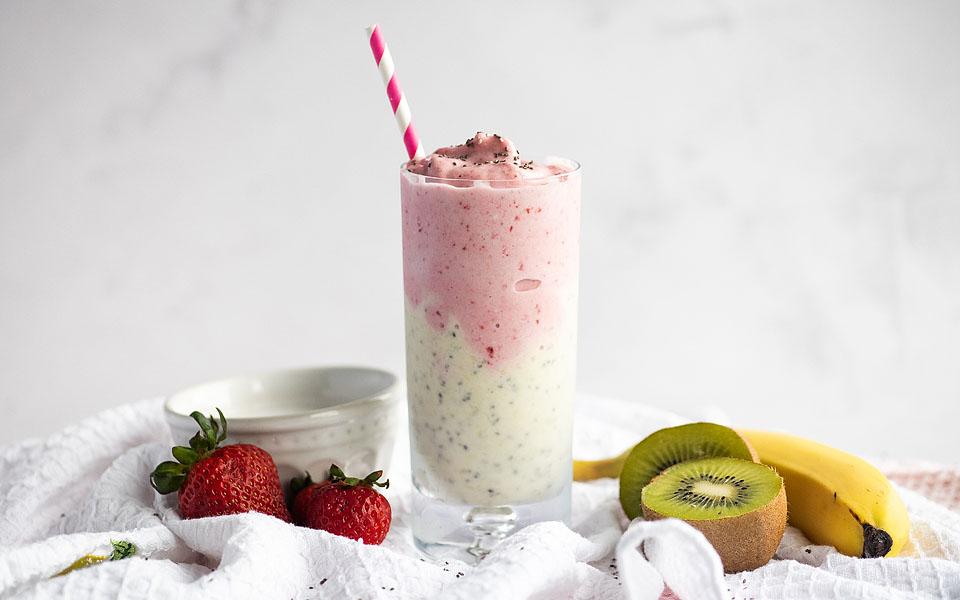 A glass of Coconut Strawberry Kiwi Smoothie with a straw next to fresh strawberries, slices of kiwi, and a banana.