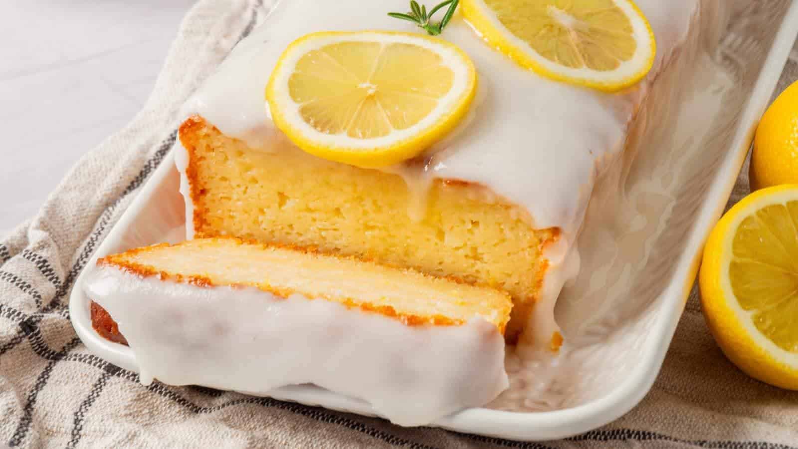 Lemon loaf cake on a white tray, topped with glaze and lemon slices, with a cut piece in the foreground and whole lemons nearby.