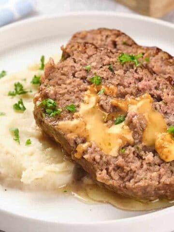 A plate with two slices of stuffed meatloaf topped with gravy, alongside mashed potatoes garnished with green herbs.