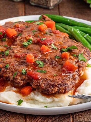 A plate of mashed potatoes topped with a Swiss Steak with some greens on the side.