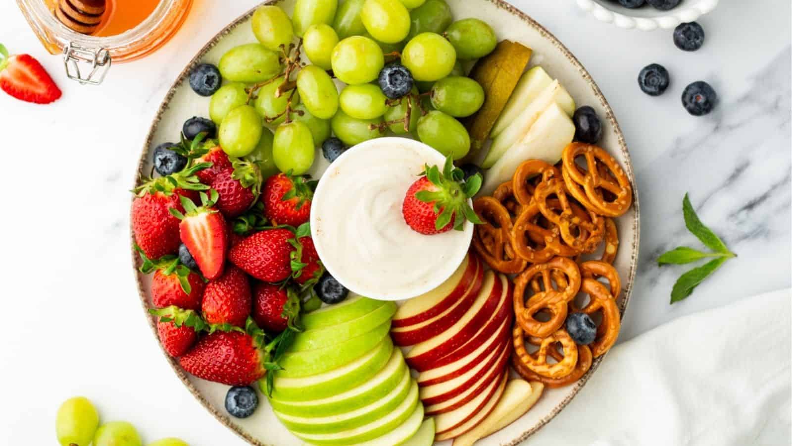 Overhead view of a bowl of yogurt fruit dip with a strawberry dipped into it, surrounded by assorted fruits and pretzels.