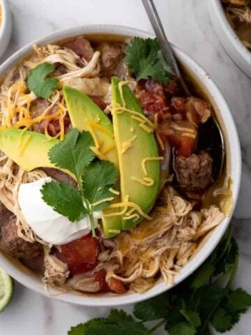 Bowl of taco soup with shredded chicken, avocado, cilantro, cheese, sour cream, and lime on a marble surface.