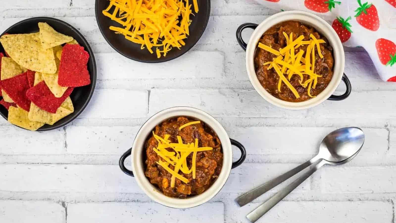Top view of Smoked Brisket Chili next to spoons, a plate of chips and shredded cheese.