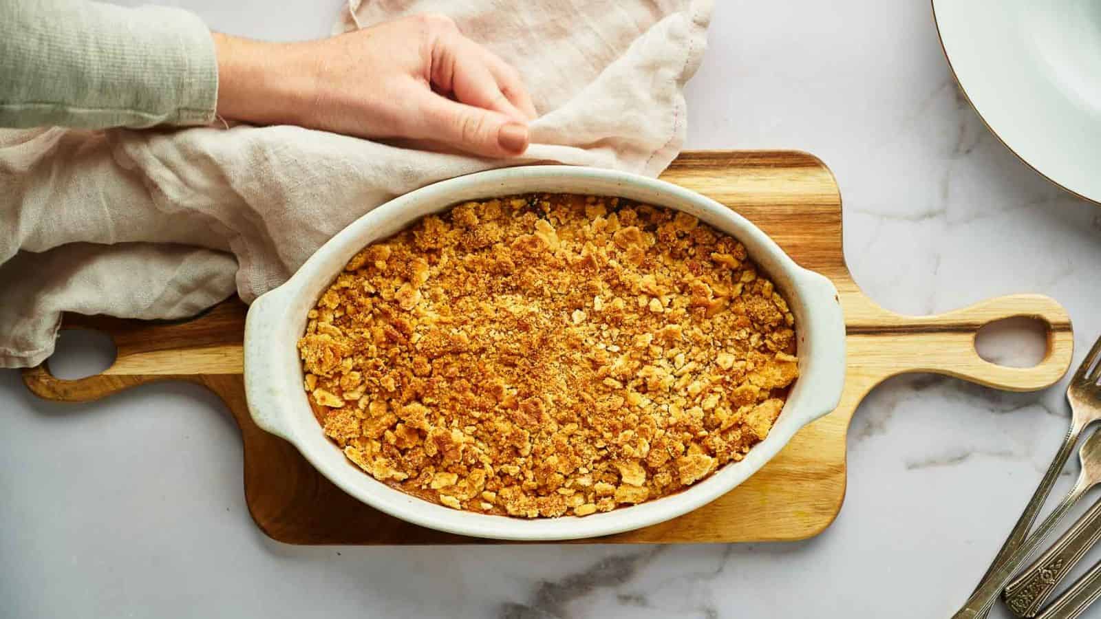 Someone holding a Yellow Squash Casserole on a wooden board with a cloth.