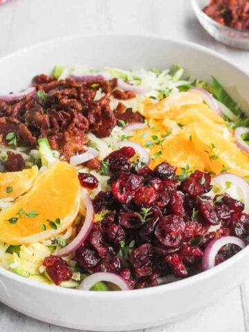 A white bowl filled with salad containing orange slices, dried cranberries, red onion, shredded greens, and bacon bits, with a fork resting inside. Small bowls of toppings and dressing nearby.
