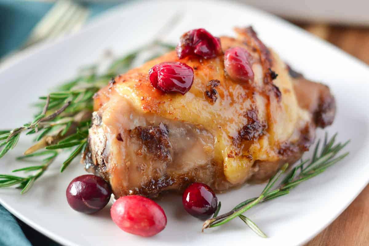 A piece of roasted chicken thighs with cranberry sauce, garnished with rosemary, rests elegantly on a white plate.