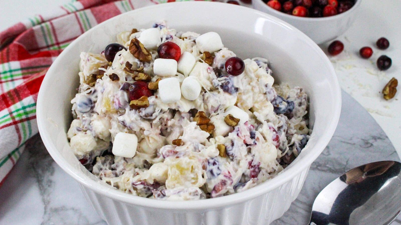 A white bowl of cranberry fluff salad topped with mini marshmallows, pecans, and cranberries on a marble surface, with a spoon and a plaid cloth nearby.
