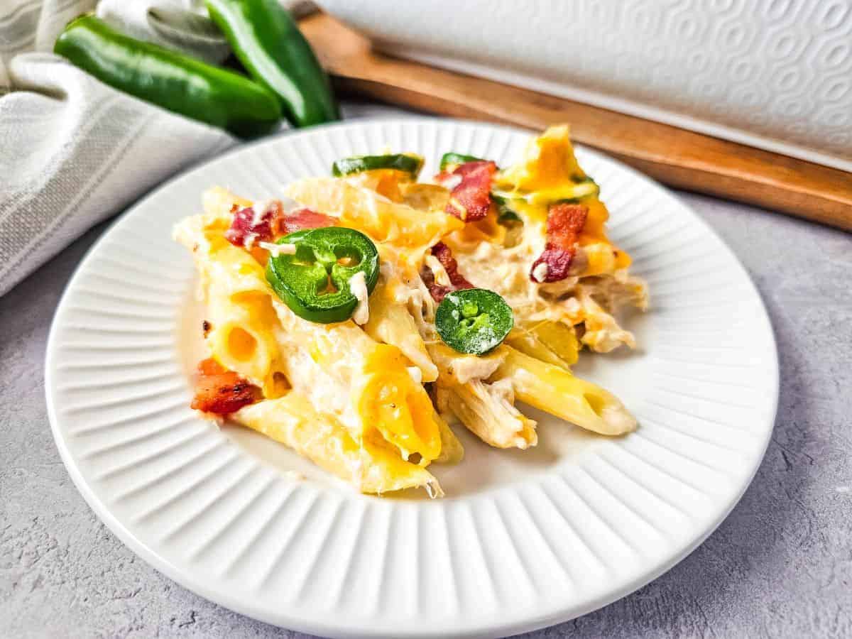 A serving spoon scoops into a baked casserole with penne pasta, cheese, jalapeños, and bacon pieces.