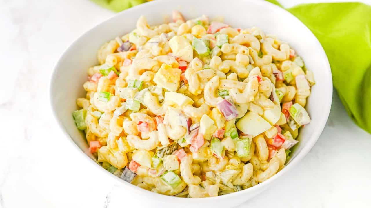 A white bowl filled with macaroni salad mixed with chopped celery, red onion, red bell pepper, and hard boiled eggs, all coated in a creamy dressing.