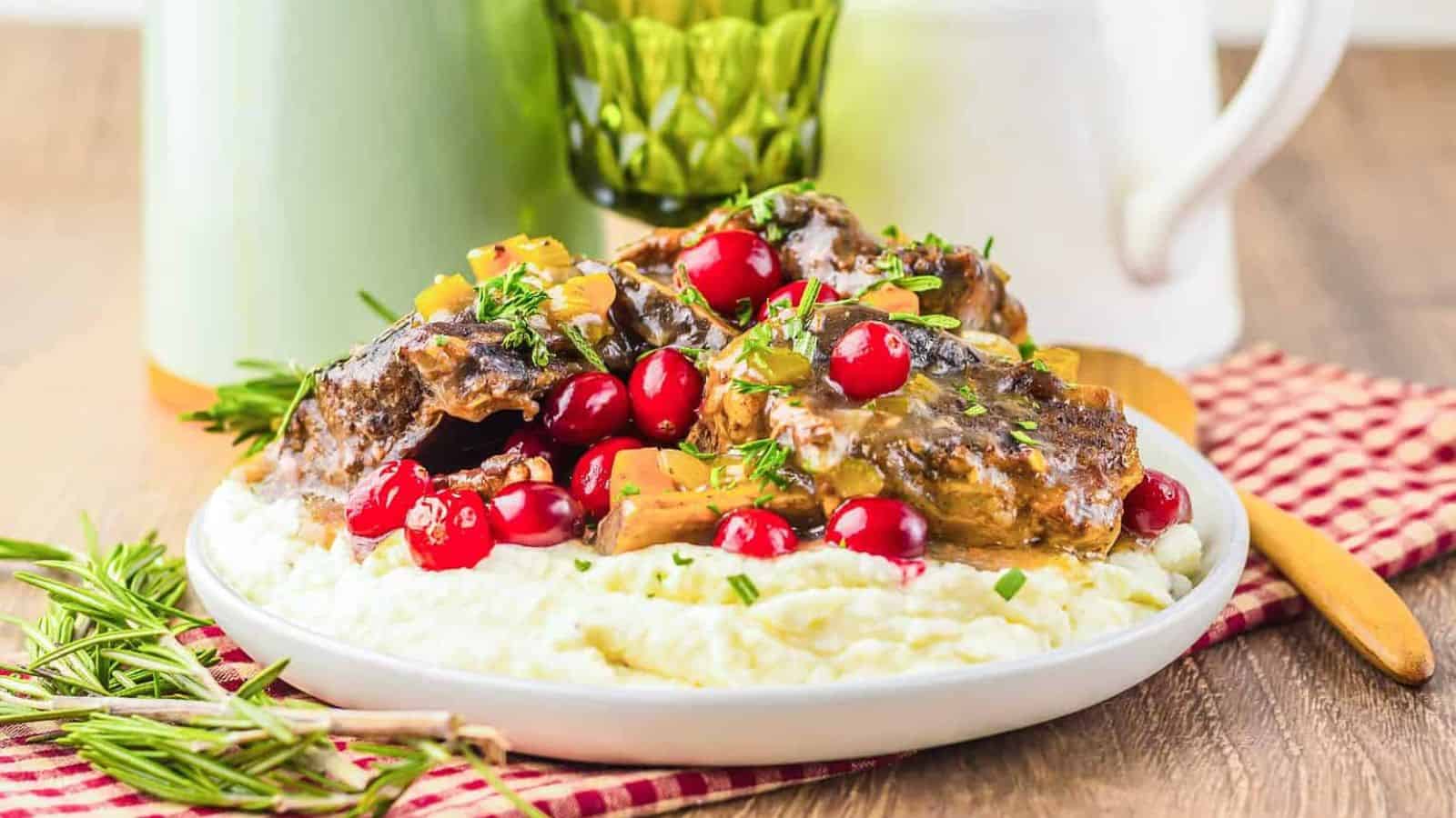 Braised meat with gravy, fresh cranberries, and herbs served over mashed potatoes on a white plate.