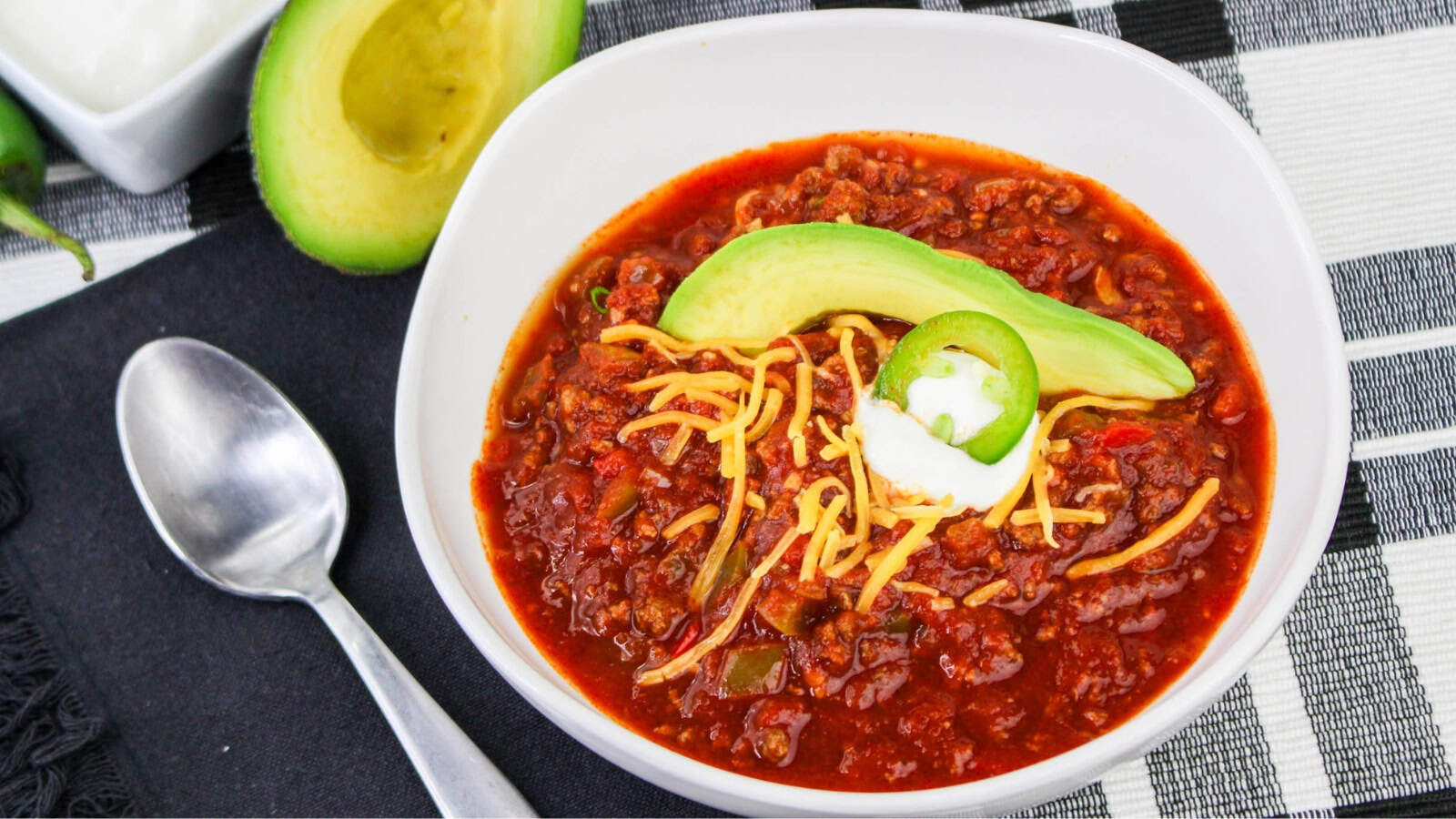 A bowl of chili topped with shredded cheese, sliced avocado, jalapeño, and a dollop of sour cream, placed on a black and white checkered cloth next to a spoon and halved avocado.