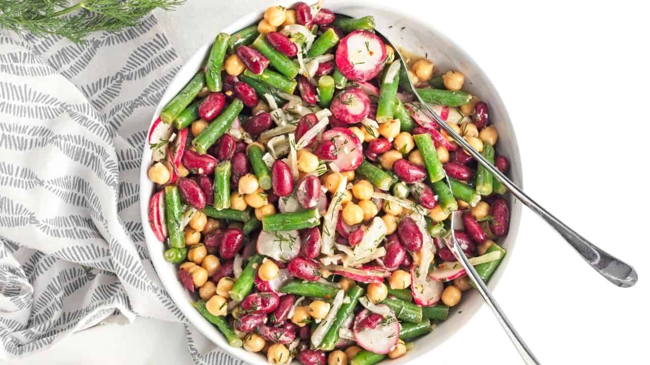 A vibrant three bean salad featuring chickpeas, kidney beans, and green beans, topped with sliced radishes and onions in a light dressing. Two metal forks rest beside a patterned cloth.