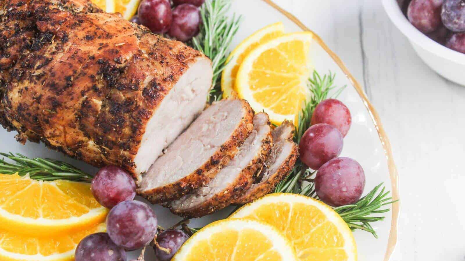 A sliced roasted pork loin garnished with rosemary, orange slices, and red grapes on a white plate, with extra grapes in a bowl nearby.