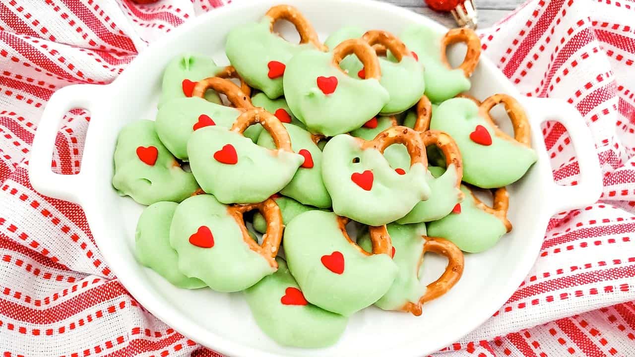A white bowl filled with pretzels coated in green frosting, each decorated with small red heart sprinkles. The bowl is placed on a red and white patterned cloth.